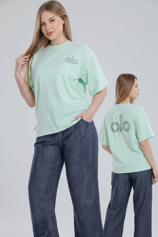 Woman wearing a light green t-shirt with 'alo' logo and blue jeans, standing in front of a white curtain with 'El Samman Group' branding.